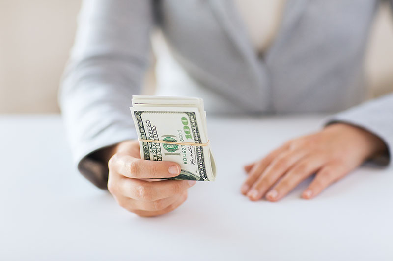 A person in a light gray suit holds out a stack of U.S. hundred-dollar bills secured with a rubber band, while their other hand rests on a white table.